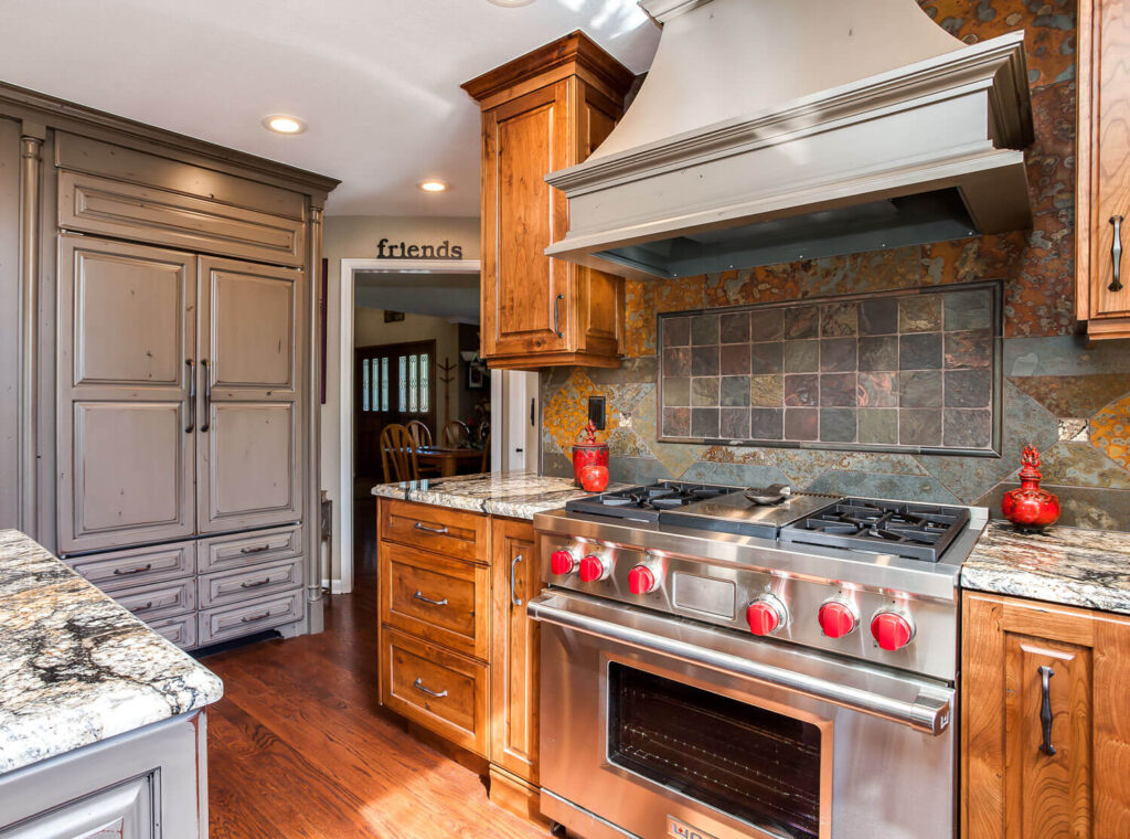 remodeled kitchen has gray distressed cabinets, large cooking hood, and unique copper backsplash JM Kitchen & Bath Design