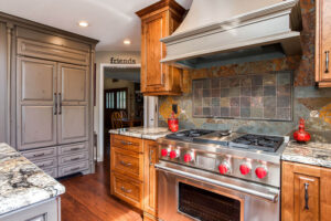 remodeled kitchen has gray distressed cabinets, large cooking hood, and unique copper backsplash JM Kitchen & Bath Design